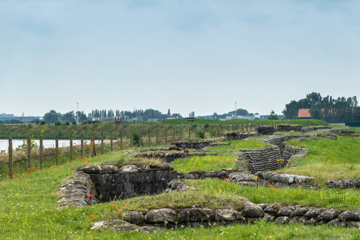 Diksmuide, Flanders, Belgium - June 19, 2019: Overview of Historic WW1 Trenches, called Dodengang along IJzer River, shows gray-brown stone-hard sandbags, green grass, red poppies. Barbed wire fence and river under blue sky.